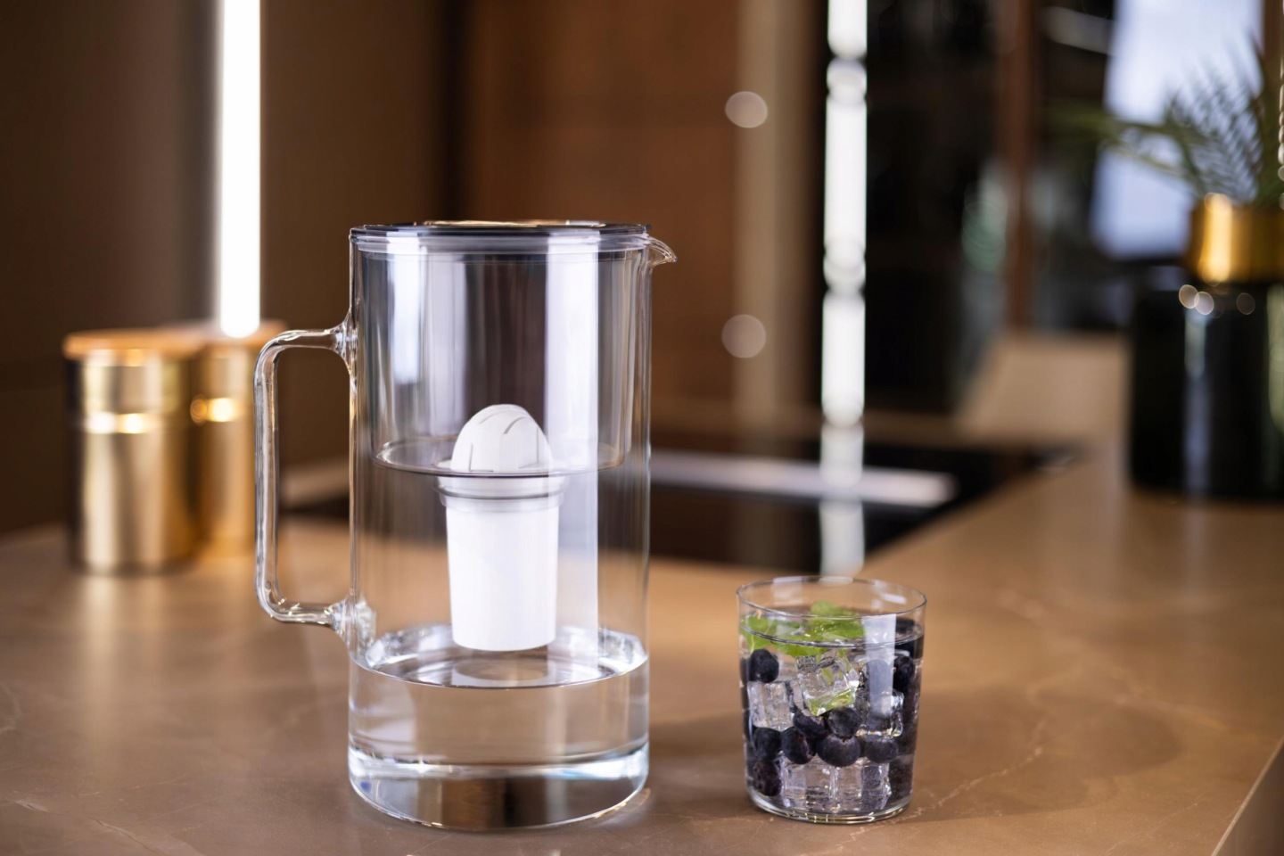 Czarny szklany dzbanek filtrujący Wessper D2 na blacie stołu obok szklanki z&nbsp;czystą wodą i&nbsp;owocami